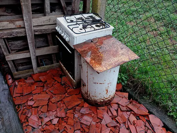 A white broken gas stove and an old washing machine stand on the old broken ceramic tiles. Unnecessary old household appliances are thrown out into the open.