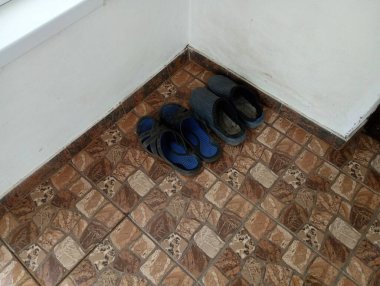 Kahverengi seramik zeminin girişinde köşedeki beyaz duvarın yanında duran iki çift lastik ayakkabı..