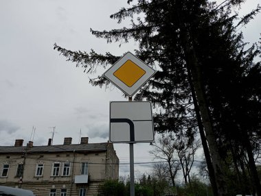 Gökyüzünün arka planına karşı ana yolu gösteren ve hareket yönünü gösteren bir işaret levhası. Beyaz arka planda sarı eşkenar dörtgen. Arka planda eski bir tuğla ev var.