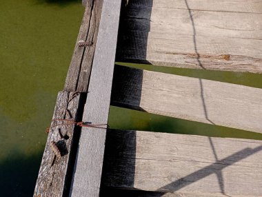 A fragment of an emergency pedestrian suspension bridge over a river with green water. A dangerous section of a wooden bridge with planks in a state of disrepair.
