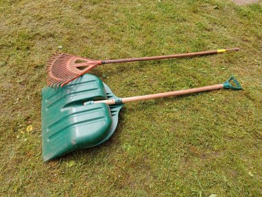Büyük yeşil bir kürek ve plastik bir turuncu tırmık biçilmiş bir çimenlikte yatıyor. Bahçe temizleme araçları.