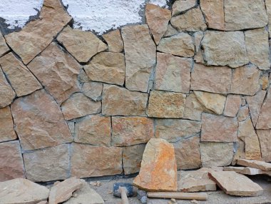 Beton bir duvarın dış cephesini doğal taşla kaplama süreci. Evin duvarına saçılmış bir sürü düz kumtaşı ve inşaat aletleri var..