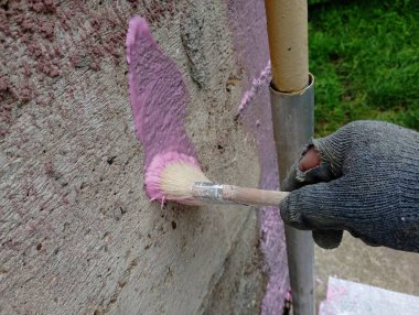 The process of priming the concrete wall of the foundation of the house with a brush by hand with a special pink concrete contact mixture. A man with a hand in a torn old glove applies primer to the wall with a brush. clipart