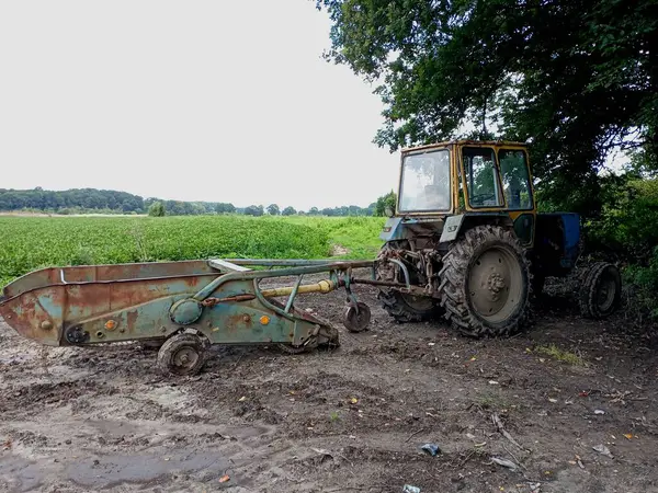 stock image An old tractor with an attached potato digger stands in the shade of forest trees on a field. The subject of agricultural technology.