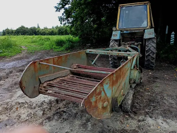 stock image An old tractor with a trailed potato digger stands in a field in the shade of trees. Agricultural machinery for digging potatoes.