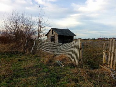 Old wooden house behind a broken wooden fence in a grassy field. Small wooden house in a field. clipart