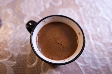 Yukarıdan, beyaz desenli masa örtüsüyle kaplı bir masanın üzerinde duran bir fincan aromatik kahve görülebilir. Kahvenin rengi köpükle kahverengidir. Bu da kalitesini ve tazeliğini gösterebilir..