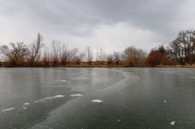 Buz yüzeyi ufka kadar uzanan donmuş bir göl ya da gölet. Sahilde çıplak ağaçlar görülebilir ve üzerlerinde ağır, bulutlu bir gökyüzü var.