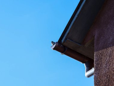 Tepeden tırnağa, kahverengi bir duvarı ve kahverengi boyanmış metal bir oluğu olan bir binanın bir kısmı görülebilir. Çatı boyunca uzanan oluklar borudan aşağı su akıtıyor..