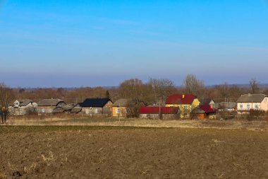 Ufuk çizgisi boyunca dizilmiş evler olan kırsal alan. Evlerin önünde, karanlık, ekili arazisi olan bir tarla uzanıyor. Arka planda yapraksız ağaçlar görülebiliyor, bu da baharın başında ya da sonbaharın sonunda olduğunu gösteriyor. Gökyüzü açık, mavi.