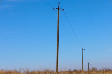 İki ahşap elektrik direği ve bir çim tarlası boyunca uzanan kablolar. Kutuplar açık mavi bir gökyüzüne karşı dizilmiştir. Bir kutup ön planda, diğeri arka planda, bir perspektif etkisi yaratıyor.