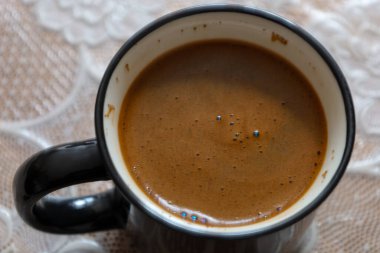 Kahveli siyah bir fincanın yukarıdan aşağı görüntüsü. İçkinin yüzeyinde küçük kabarcıklar görülebilir. Bardak desenli bir kumaşın üzerinde duruyor..