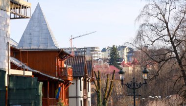 Fotoğraf, hem antik hem de modern mimarinin unsurlarıyla kentsel bir manzara yakalıyor. Ön planda, bir şatoya benzeyen ahşap süslemeli ve sivri uçlu çatılı bir binanın bir parçası görülebilir..
