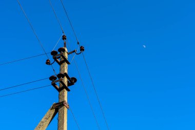 Fotoğraf, parlak mavi gökyüzünün zeminine yalıtkan ve kablolar döşenmiş beton bir elektrik direğini tasvir ediyor. Çerçevenin sağ üst köşesinde küçük bir ay görünür..