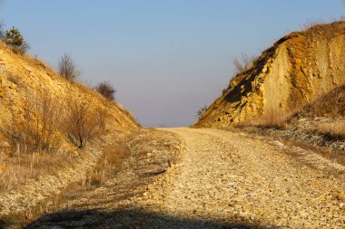 Fotoğraf iki tepenin arasından geçen çakıllı bir yolu tasvir ediyor. Tepelerin sarımsı kahverengi bir tonu ve düz olmayan bir yüzeyi vardır. İnce çalılar ve dalları olan ağaçlar tepelerde yetişir..