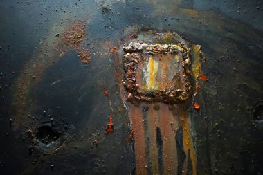 Siyah arkaplanda düz olmayan kenarları ve pas çizgileri olan kare bir kaynak izi görülebilir. Yüzeyde başka aşınma ve hasar izleri de var..