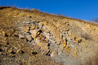 Güneşli bir gün. Ön planda, farklı renk ve dokulardan oluşan kaya katmanlarının görülebildiği, çıplak toprakla kaplı bir yamaç vardır. Taşlar yüzeye saçılmış..