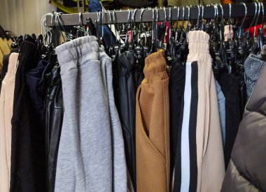 Mağazadaki elbise askılarının yakın çekimleri. Metal askılarda asılı çeşitli pantolonlar görülebilir. Pantolonun farklı renkleri ve malzemeleri var.