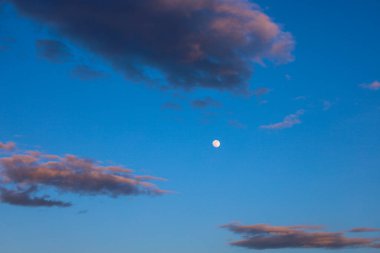 Fotoğraf, batan güneşin son ışınlarıyla aydınlanan birkaç kabarık bulutlu mavi bir gökyüzünü gösteriyor. Çerçevenin ortasında küçük bir ay görünür..