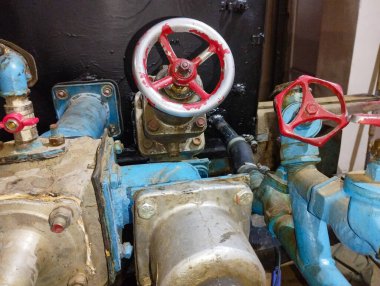 Ön planda kırmızı ve gümüş saplı iki büyük metal valf görülebilir. Borular ve diğer ekipman elementleri mavi ve gri, aşınma ve aşınma izleri var..