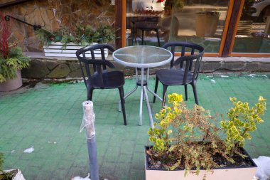 Fotoğrafta açık bir kafe, yuvarlak bir masa ve iki sandalye var. Masanın cam bir üstü ve metal bacakları var. Sandalyeler plastik, siyah renkli..