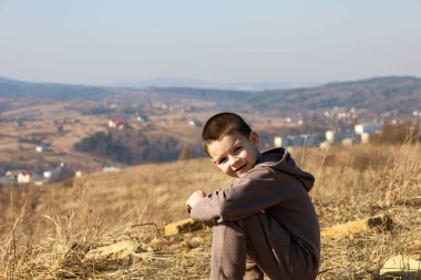 Genç bir çocuk, bir tepede, kuru çimlerin arasında oturuyor, kameraya gülümsüyor. Arka plan, dağınık evleri ve yuvarlanan tepeleri olan kırsal bir manzaranın panoramik görüntüsünü ortaya çıkarıyor..