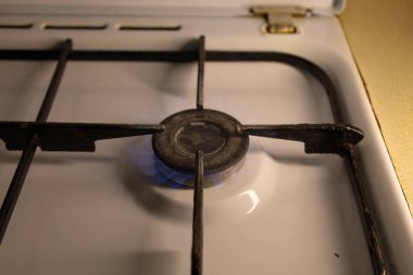 Fotoğraf, yanan mavi bir alevle bir gaz sobasının yakın çekimini gösteriyor. Sobanın beyaz bir diş minesi ve dökme demir ızgaraları var..