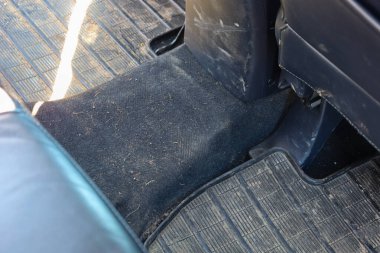 Bu fotoğraf kirli bir arabanın içini gösteriyor. Lastik paspaslar ve kumaş kaplı. Lastik paspaslar siyah, tırtıklı deseni var ve kumaş zemin kaplaması koyu gri ya da siyah..