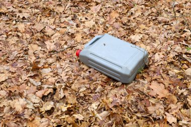 Bu fotoğraf, ormandaki dökülen yaprakların arasına atılmış plastik bir teneke kutuyu gösteriyor. Kutu gri ve kırmızı kapaklı. Kahverengi yapraklarla bir kontrast oluşturuyor..