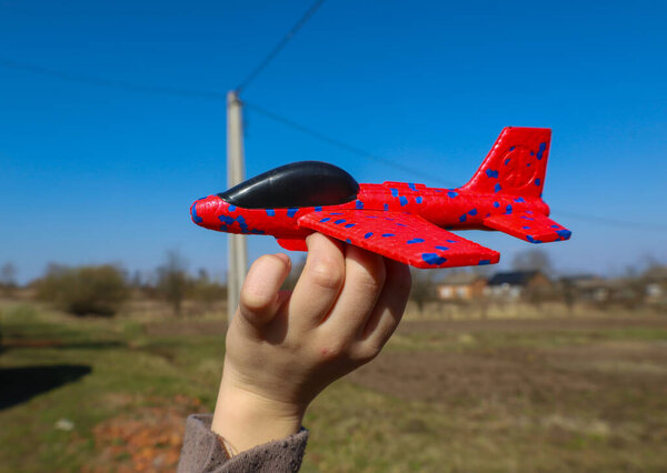 A close-up photograph shows a child's hand holding a red toy airplane against a backdrop of a blue sky and rural landscape. The airplane has a black cabin and blue spots on its wings.