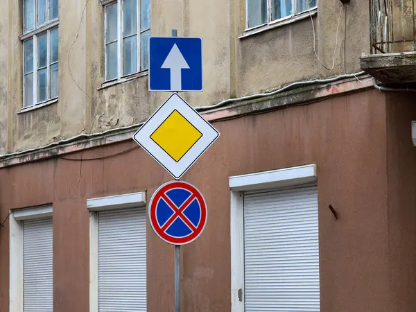 Fotoğrafta, birbiri üstüne dizilmiş üç yol işareti var. Bir direğin üzerinde, patenli panjurlu bir binanın arka planına karşı..