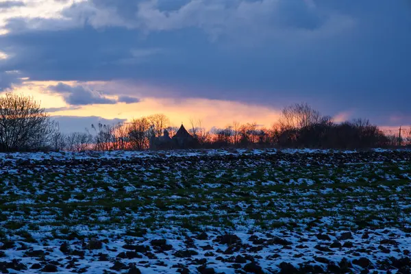 Fotoğraf günbatımında kırsal bir manzarayı gösteriyor. Ön planda, koyu toprak ve yeşil lahanalarla kaplı karla kaplı bir alan görülebilir. Arka planda, bir sıra ağaç bir kilisenin siluetini saklıyor..