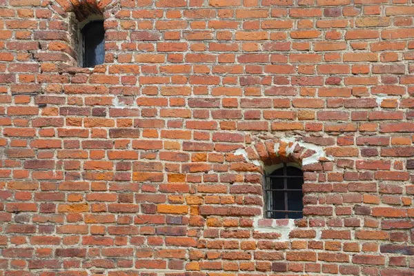 Fotoğraf iki küçük pencereli kırmızı tuğladan yapılmış bir tuğla duvarı gösteriyor. Tuğlanın çeşitli tonlarında kırmızı ve doku var. Tuğlaların arasındaki dikişler duvarda görünüyor..