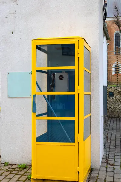 Fotoğraf bir binanın yanındaki caddede bulunan sarı bir telefon kulübesini gösteriyor. Kabinin şeffaf cam panelleri ve sarı metal çerçevesi var. Kabinin içinde, bir telefon aygıtı görünüyor..