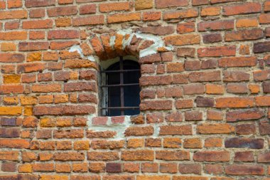 Fotoğraf, küçük pencereli kırmızı tuğladan yapılmış bir tuğla duvarı gösteriyor. Tuğlanın çeşitli tonlarında kırmızı ve doku var. Tuğlaların arasındaki dikişler duvarda görünüyor..