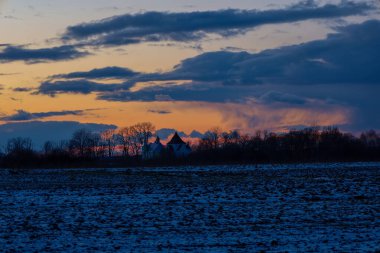Fotoğraf, ufukta görünen bir kilisenin olduğu kırsal bir manzarayı gösteriyor. Ön plandaki alan kısmen karla kaplı. Kilisenin beyaz duvarları ve karanlık bir çatısı var. Şey...
