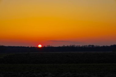 Fotoğraf bir tarlanın üzerinde gün batımını gösteriyor. Güneş ufuk çizgisinin arkasında batıyor, gökyüzünde parlak turuncu ve sarı bir eğim yaratıyor..