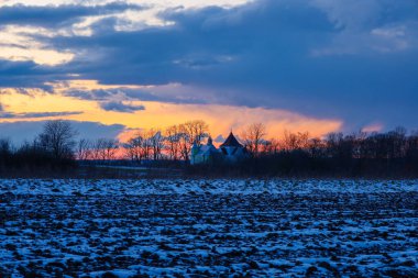 Kış kırsalında gün batımında. Ön planda, donmuş toprak kümeleriyle kaplı karla kaplı bir tarla. Tarlanın arkasında, küçük bir kilisenin ya da kulesi ve sivri bir çatısı olan eski bir binanın silueti görülebilir, çıplak ağaçlarla çevrili..
