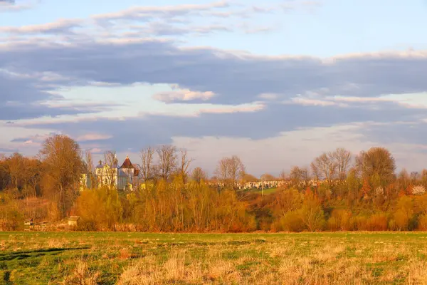 İlk yeşil yaprakları olan yapraksız ağaçlar ve ağaçlar kiliseyi beyaz duvarlar ve kırmızı çatılarla çevreler. Mezarları kilisenin arkasındaki tepede görünüyor. Huzurlu akşam manzarası, Ukrayna ruhaniliği, kırsal alan.