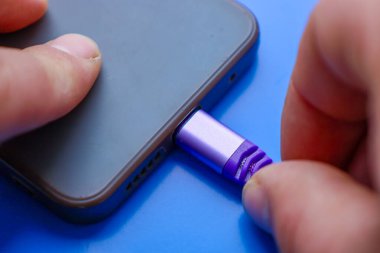 Mor bir kabloyu gri bir akıllı telefonun şarj portuna bağlama işlemine yakın çekim. Parmaklar dikkatlice konnektörü yerleştirirken görünür..