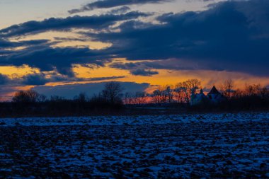 Önünde karla kaplı bir tarla, ufukta ağaçların siluetleri ve dramatik turuncu ve mavi gün batımının altında bir bina olan kış akşamı manzarası..