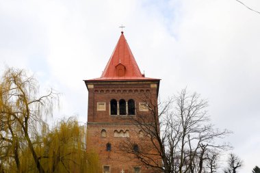 Görkemli bir tuğla kule yükseliyor, kırmızı çatısı soluk gökyüzüne şiddetle zıt düşüyor. Kemerli pencereler ön cepheyi süslüyor, tarihsel cazibesine ekliyor..