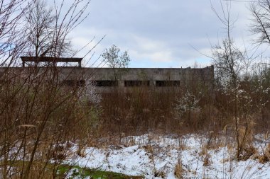 Boş pencere açıklıkları olan terk edilmiş beton bir bina, kasvetli bir kış gününde çalılıkların ve karla kaplı çimenlerin arasından görülebilir..