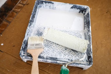 Beyaz boyalı bir boya tepsisinin üst görüntüsü, içinde boya silindiri var. Kartonun yanında, tahta saplı ve boyayla boyanmış kıllı bir boya fırçası var..