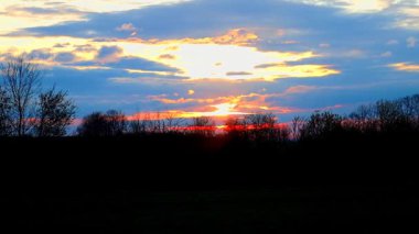 Büyüleyici bir günbatımı, ağaçların tepelerini sıcacık ışığıyla kucaklıyor. Güneş ufuk çizgisinin gerisine doğru yavaşça iniyor, ağaçların siluetleri alevler içindeki gökyüzüne karşı karanlık bir zıtlık olarak göze çarpıyor..