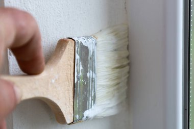 Beyaz boyalı fırçayı tutan bir elin yakın çekimi ve bir duvarla pencere çerçevesi arasındaki köşeyi boyamak. Taze boyayla kaplı fırçanın kılları, ahşap bir sapı ve metal bir gelincik görülebilir..