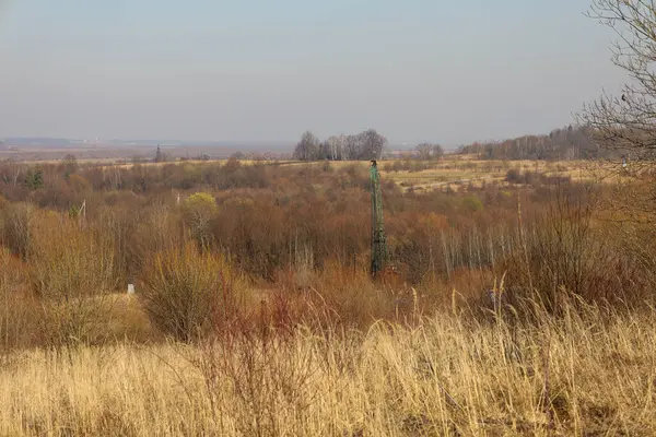 Arka planda, soluk, puslu bir gökyüzünün altında tarlaları ve ağaçları olan bir düzlük görülebilir. Uzun yeşil bir metal yapı, muhtemelen sulama sisteminin bir parçası veya bir kule, merkezde duruyor..