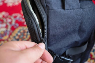 Biri koyu mavi bir sırt çantasının cebini kapatıyor. Fermuarı tutan parmakları olan bir elin parçası görülebilir. Arka planda, kırmızı ve altın rengi bulanık bir desen var..