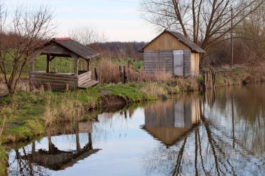 Küçük ahşap bir çardağı ve sakin bir göletin kıyısında evi olan rahat bir kırsal alan. Binalar ve çevreleyen yapraksız ağaçlar durgun suya çok güzel yansıyor..