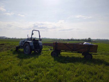 Ukrayna 'da güneşli bir bahar gününde yeşil bir tarlada açık gri bir traktör duruyor. Traktör solda yer alıyor ve sağda duran tahta kenarlı bir römorka bağlı..